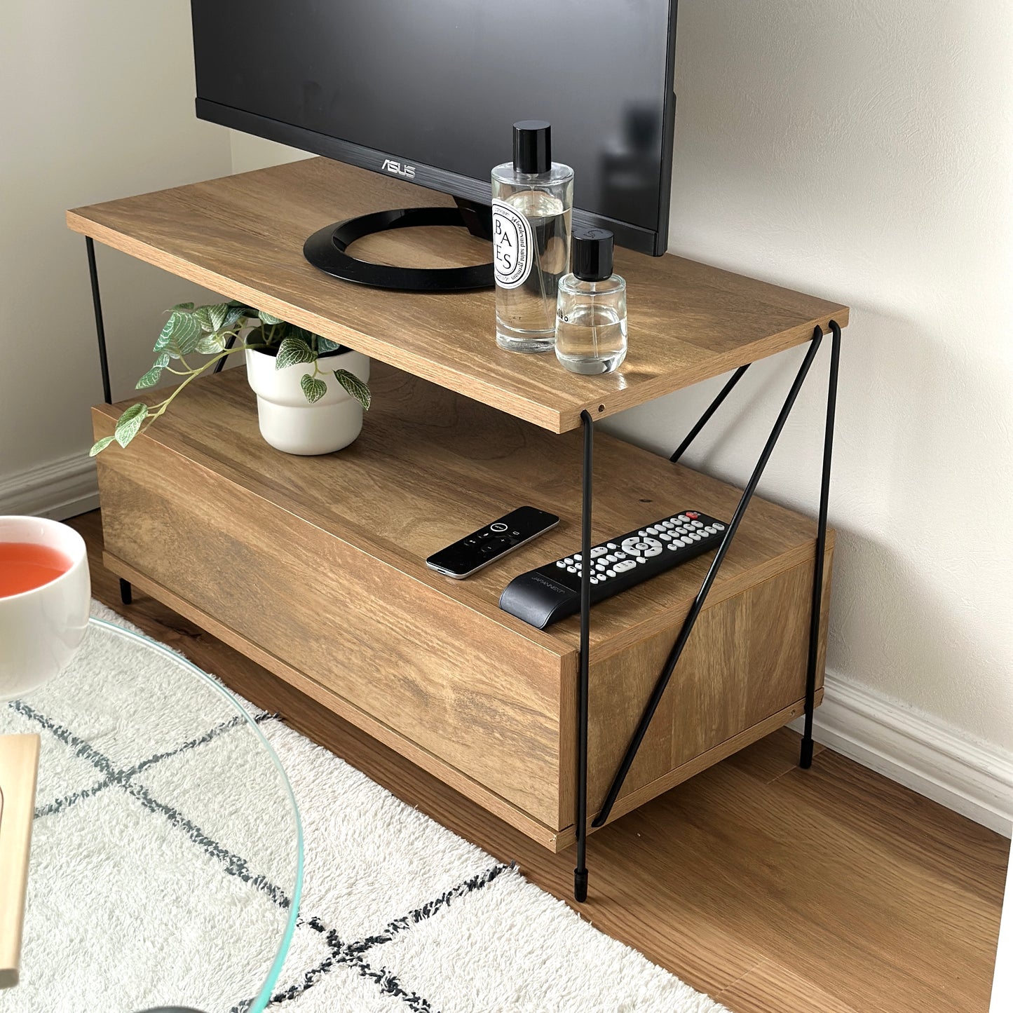 木目のウッドと細いブラックフレームのテレビボードに香水と植物、リモコンを置いている様子
